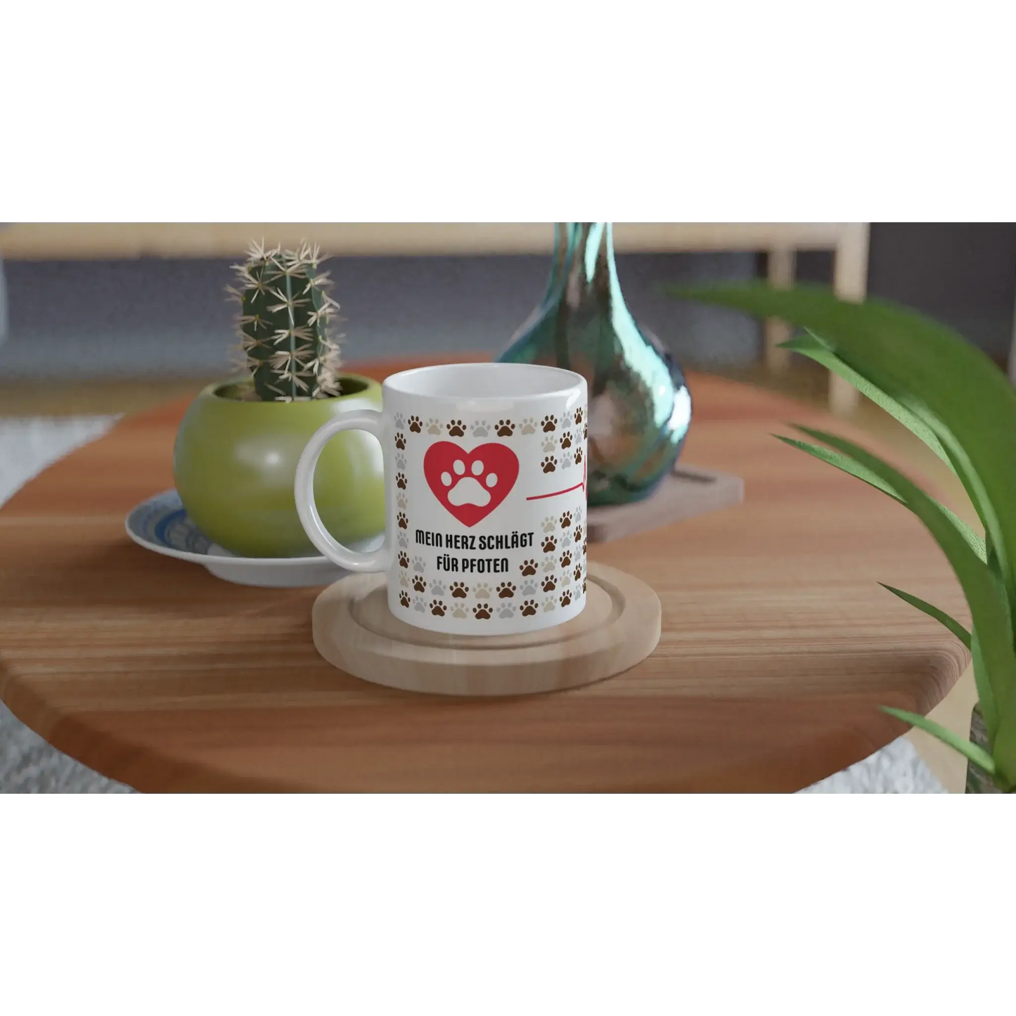 Die Tasse „Mein Herz schlägt für Pfoten“ von Tasse mit Herz, mit Herz- und Pfotenmotiv und deutschem Text, steht mit einem Untersetzer auf einem Holztisch. Daneben vervollständigen ein kleiner Kaktus in einem grünen Topf und eine Glasvase dieses charmante Geschenkset für Tierfreunde.