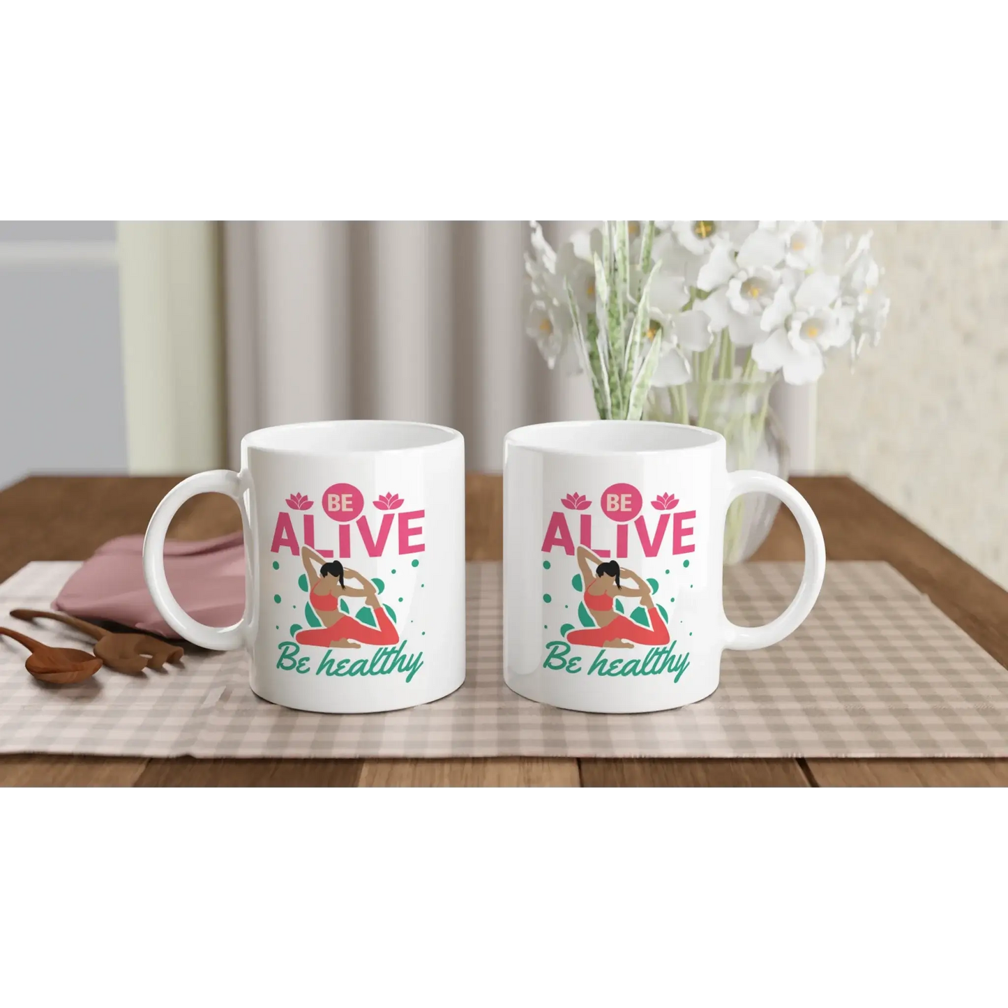 Zwei weiße „Achtsamkeits-Tasse ‚Be Alive, Be Healthy‘“ von Tasse mit Herz stehen auf einem karierten Tischset. Jede Tasse zeigt eine Yoga-Frau und den Text. Im Hintergrund stehen eine weiße Vase mit Blumen, ein rosa Buch und Holzutensilien – ideal als Yoga-Geschenk oder Tasse Achtsamkeit.
