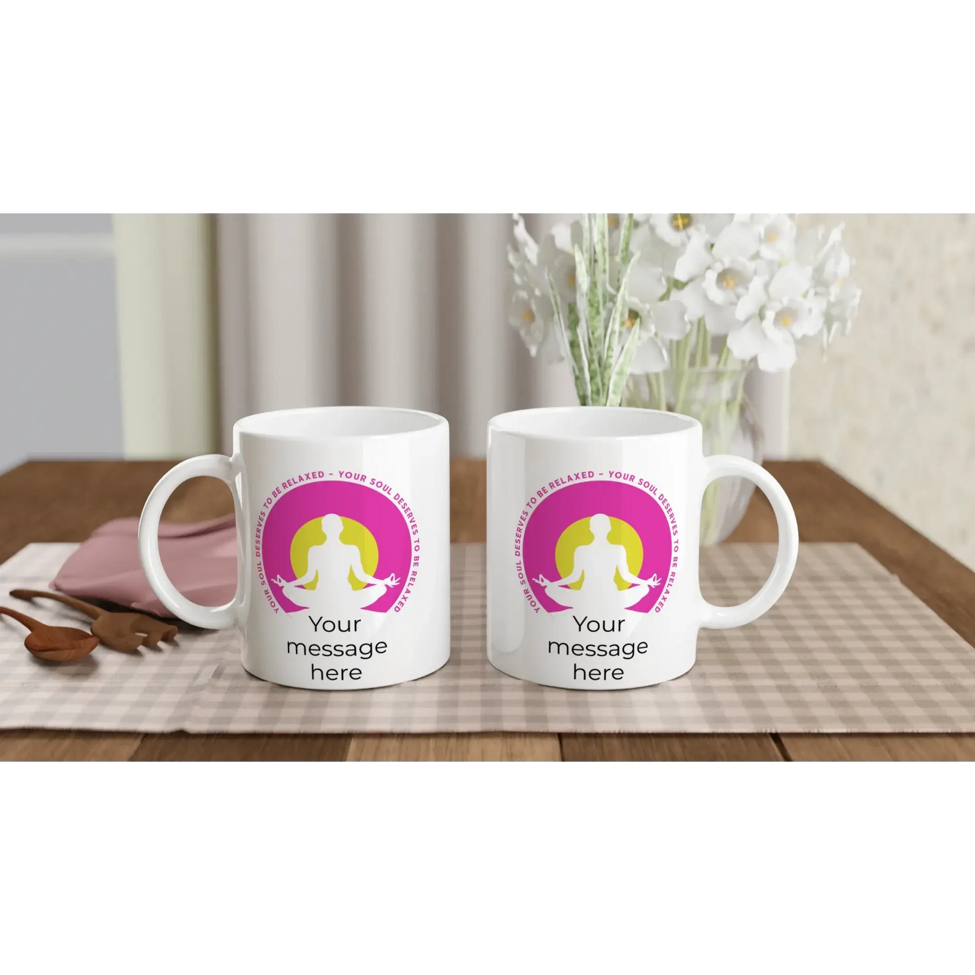 Zwei weiße Tassen von Tasse mit Herz mit dem pinkfarbenen Motiv einer meditierenden Person und dem Satz „Deine Seele verdient Entspannung“ stehen auf einem Tisch neben einer pinkfarbenen Kosmetiktasche und Holzbesteck. Weiße Blumen tragen zur friedlichen Atmosphäre bei.