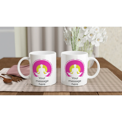 Zwei weiße Tassen von Tasse mit Herz mit dem pinkfarbenen Motiv einer meditierenden Person und dem Satz „Deine Seele verdient Entspannung“ stehen auf einem Tisch neben einer pinkfarbenen Kosmetiktasche und Holzbesteck. Weiße Blumen tragen zur friedlichen Atmosphäre bei.