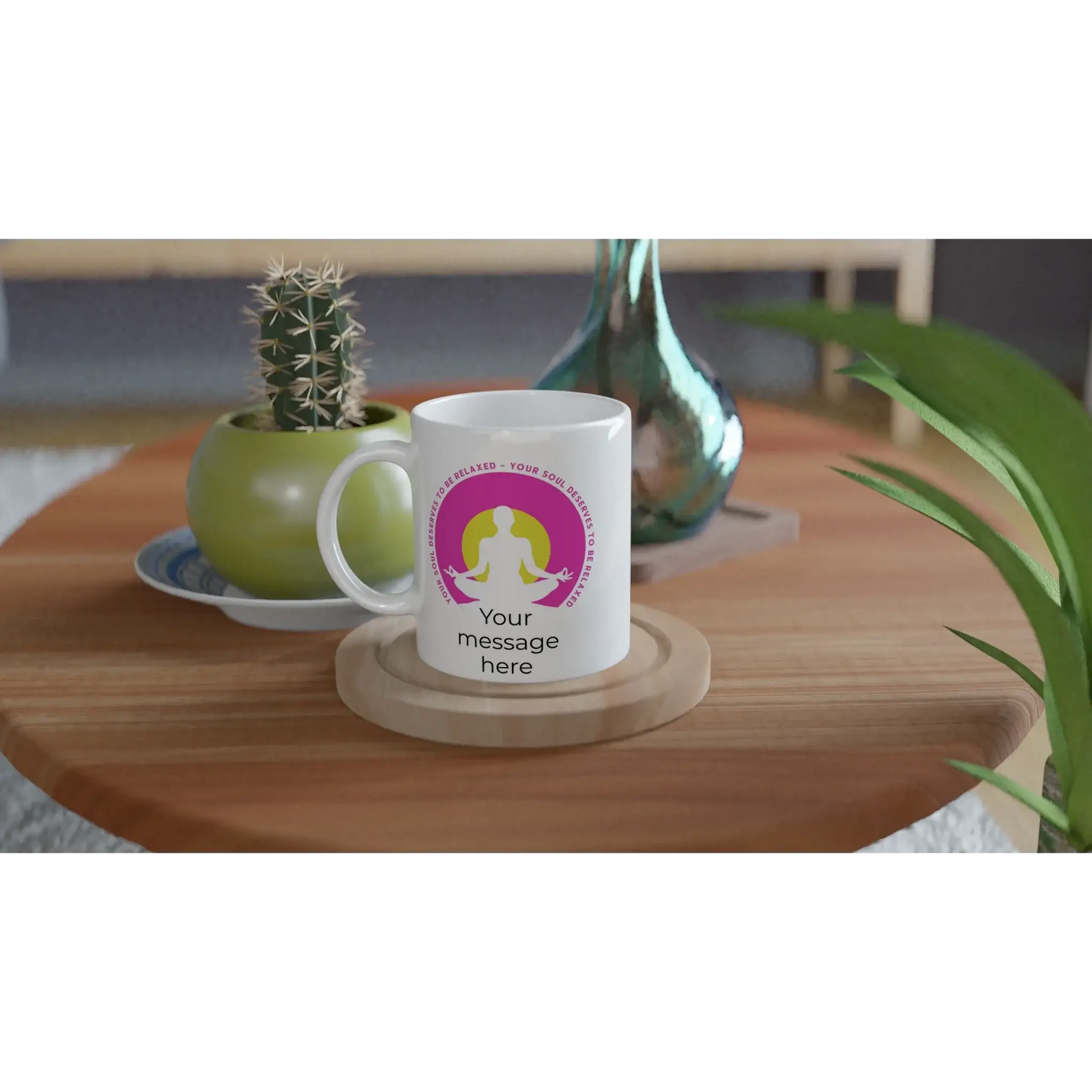 Eine weiße Tasse auf einem Holztisch zeigt die Grafik einer meditierenden Person in einem rosa Kreis, der Achtsamkeit und Harmonie symbolisiert. Der Text lautet: „Deine Seele verdient Entspannung.“ Neben der personalisierbaren Yoga-Tasse von Tasse mit Herz steht ein kleiner Kaktus in einem grünen Topf und einer blauen Vase.