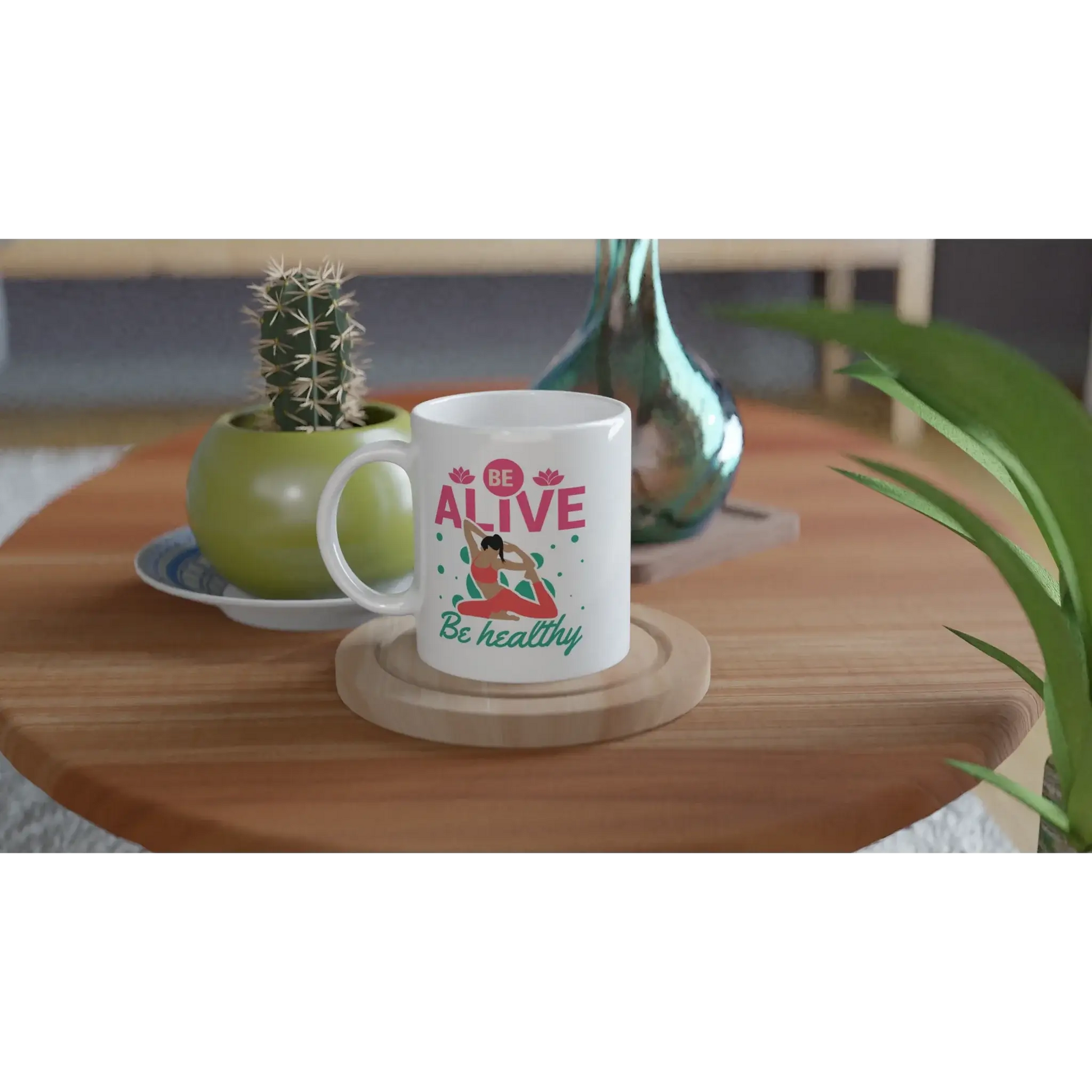 Die Achtsamkeits-Tasse „Be Alive, Be Healthy“ von Tasse mit Herz steht auf einem Holztisch, begleitet von einem Kaktus im grünen Topf und einer dekorativen Glasflasche – eine perfekte, ruhige Umgebung für jeden Liebhaber von Gesundheitsaccessoires.