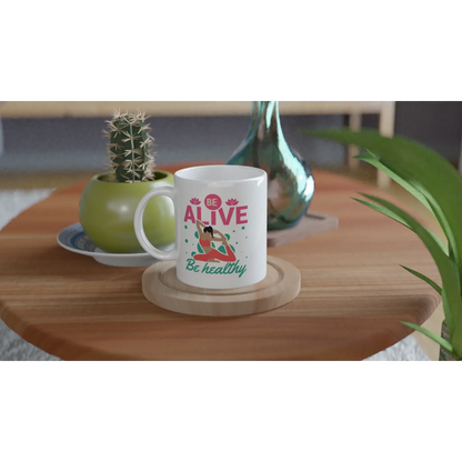 Die Achtsamkeits-Tasse „Be Alive, Be Healthy“ von Tasse mit Herz steht auf einem Holztisch, begleitet von einem Kaktus im grünen Topf und einer dekorativen Glasflasche – eine perfekte, ruhige Umgebung für jeden Liebhaber von Gesundheitsaccessoires.
