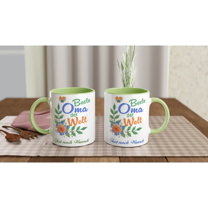 Two Tasse mit Herz mugs with green handles rest on a checkered tablecloth, each featuring Beste Oma der Welt in colorful text with floral designs. These charming Tassen are the perfect Geschenk. The background reveals part of a wall and curtain.