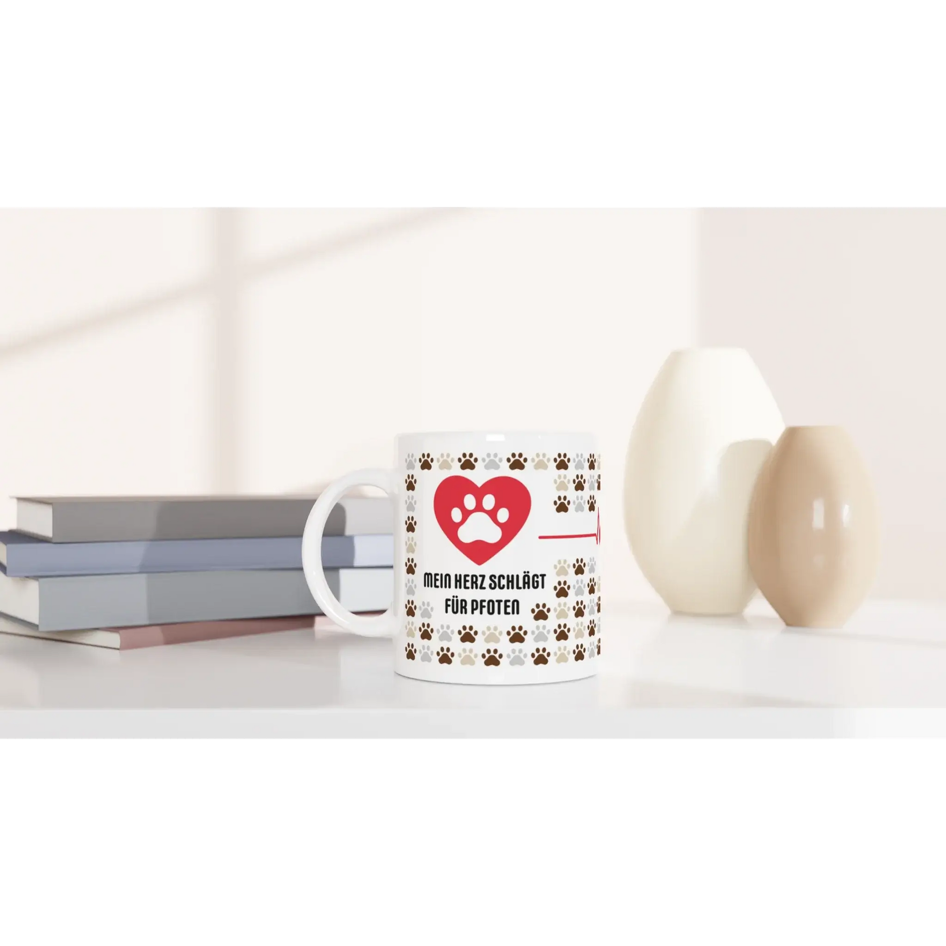 Die Tasse mit Herz aus der Produktlinie „Mein Herz schlägt für Pfoten“ besteht aus einer weißen Tasse mit Pfotenabdrücken, platziert neben Büchern und Vasen. Ideal als Geschenk für Tierfreunde, die sanfte Beleuchtung unterstreicht die gemütliche Atmosphäre.