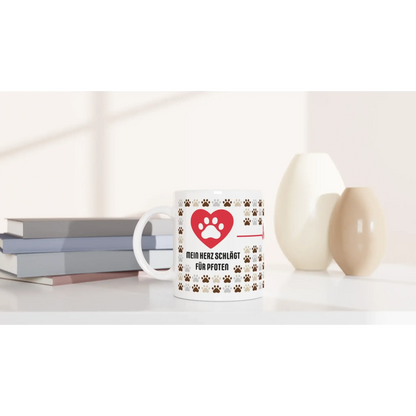 Die Tasse mit Herz aus der Produktlinie „Mein Herz schlägt für Pfoten“ besteht aus einer weißen Tasse mit Pfotenabdrücken, platziert neben Büchern und Vasen. Ideal als Geschenk für Tierfreunde, die sanfte Beleuchtung unterstreicht die gemütliche Atmosphäre.