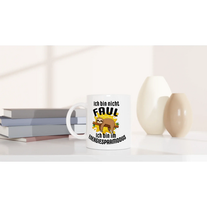 Der „Humorvoller Kaffeebecher“ von Tasse mit Herz enthält einen deutschen Text und ein Cartoon-Faultier mit der Aufschrift „Ich bin nicht faul, ich bin im Energiesparmodus“. Perfekt für einen entspannten Lebensstil, steht er auf dem Tisch neben gestapelten Büchern und dekorativen Vasen.