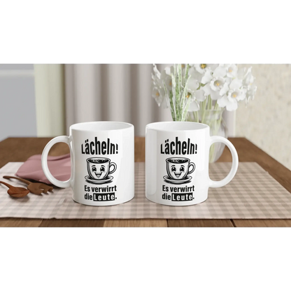 Auf einer karierten Tischdecke stehen zwei weiße Tassen von „Tasse mit Herz“ mit Cartoon-Gesichtern und dem deutschen Text „Lächeln! Es ist verrückt die Leute“, die eine humorvolle Tasse ergänzen. Weiße Blumen und ein rosa Notizbuch sorgen für Charme – ein perfektes Geschenk für Fans des Frecher-Spruchs mit dem Spruch „☕️ Lächeln! Verwirre die Welt.“ 📚🌸.