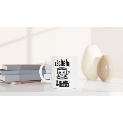 Die Tasse „Lächeln! Verwirre die Welt“ von Tasse mit Herz zeigt einen lustigen Spruch und eine lächelnde Teetassengrafik auf Weiß. Zusammen mit Büchern und Vasen ist es das perfekte Geschenk für humorvolle Tassenliebhaber.