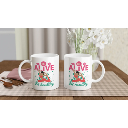 Zwei weiße „Achtsamkeits-Tasse ‚Be Alive, Be Healthy‘“ von Tasse mit Herz stehen auf einem karierten Tischset. Jede Tasse zeigt eine Yoga-Frau und den Text. Im Hintergrund stehen eine weiße Vase mit Blumen, ein rosa Buch und Holzutensilien – ideal als Yoga-Geschenk oder Tasse Achtsamkeit.