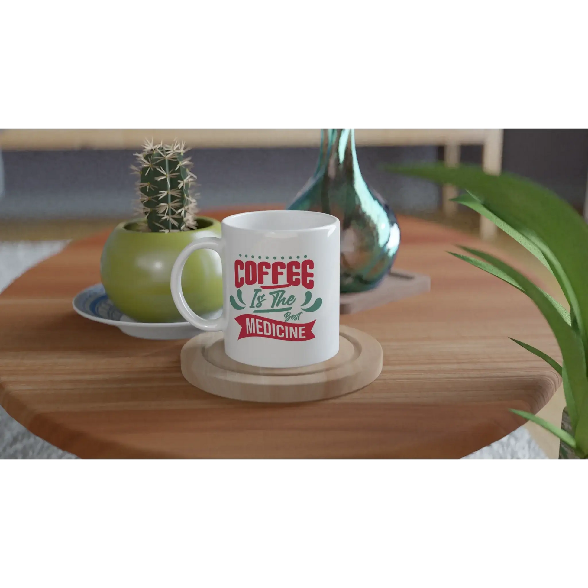 Eine Tasse mit Herz mit der Aufschrift „Kaffee ist die beste Medizin“ steht auf einem Holztisch. Daneben steht ein grüner Keramiktopf mit einem Kaktus, ergänzt durch eine reflektierende blaugrüne Vase. Dieses Ensemble ergibt ein ideales Geschenk für Kaffeefans mit Humor.