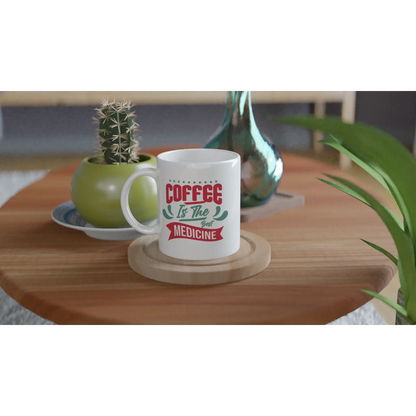 Eine Tasse mit Herz mit der Aufschrift „Kaffee ist die beste Medizin“ steht auf einem Holztisch. Daneben steht ein grüner Keramiktopf mit einem Kaktus, ergänzt durch eine reflektierende blaugrüne Vase. Dieses Ensemble ergibt ein ideales Geschenk für Kaffeefans mit Humor.