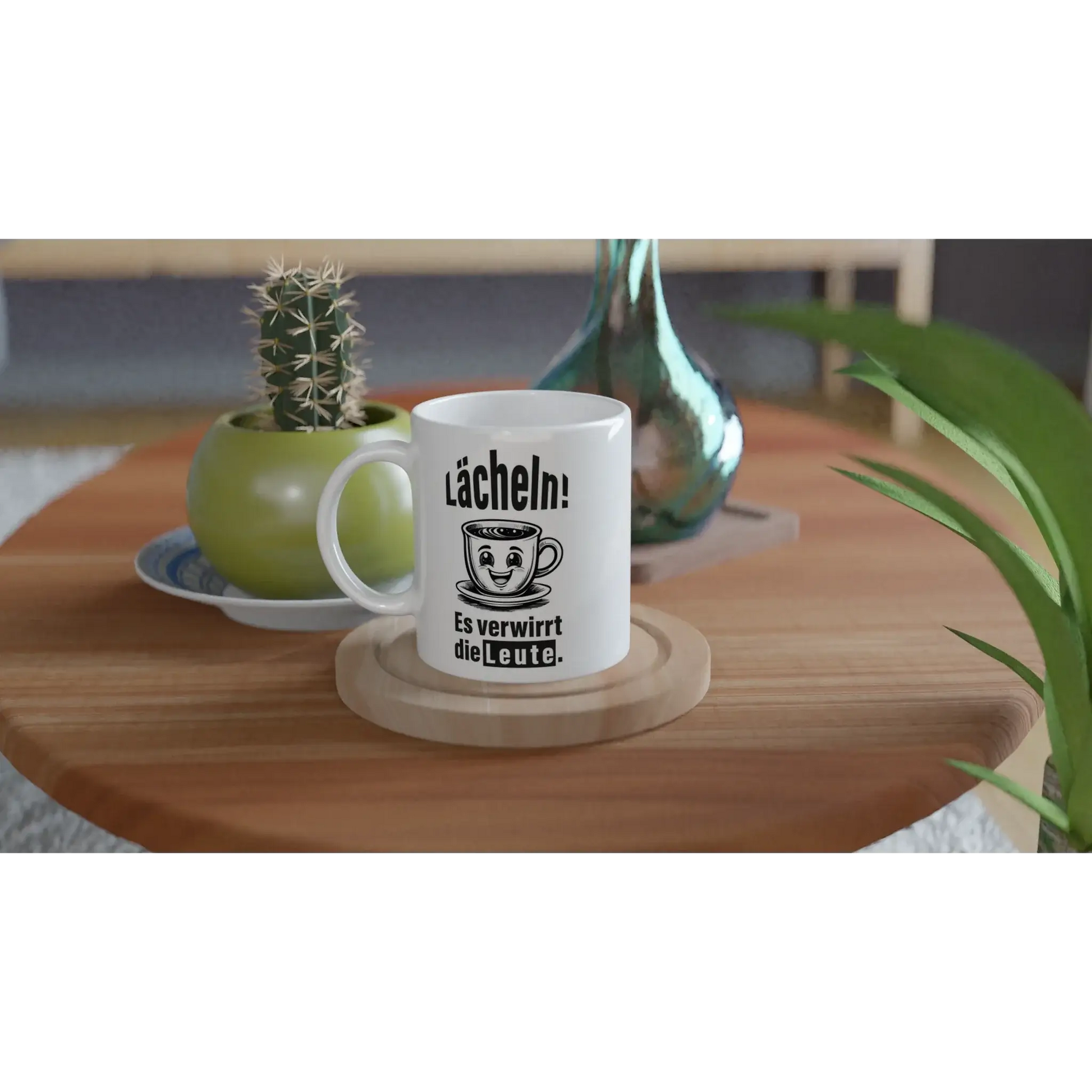 Das „☕️ Lächeln! Verwirre die Welt – Deine humorvolle Statement-Tasse!“ von Tasse mit Herz steht auf einem Holztisch, daneben ein kleiner Kaktus in einem grünen Topf und eine blau-grüne Vase mit grünem Blatt – ideal als frecher Spruch-Geschenkidee. 😂.