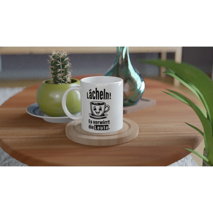 Das „☕️ Lächeln! Verwirre die Welt – Deine humorvolle Statement-Tasse!“ von Tasse mit Herz steht auf einem Holztisch, daneben ein kleiner Kaktus in einem grünen Topf und eine blau-grüne Vase mit grünem Blatt – ideal als frecher Spruch-Geschenkidee. 😂.